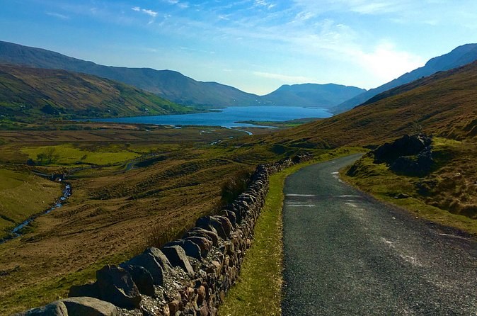 A narrow road beside a dry stone wall, running down a valley towards a long lough